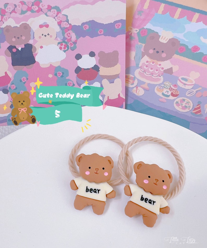 May include: Two brown teddy bear hair ties with beige rope-like bands. Each bear wears a white shirt with the word "bear" printed on it. The background includes a pink and purple illustration with teddy bear characters and the text "Cute Teddy Bear".