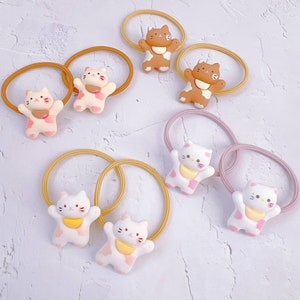 Puede incluir: Colecci&oacute;n de gomas para el pelo con peque&ntilde;as figuras de gatos en 3D en varios colores. Los gatos tienen detalles rosas, marrones y blancos, con bandas el&aacute;sticas a juego en tonos rosas y amarillos. El fondo es una superficie clara y texturizada.