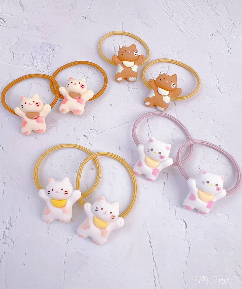 Puede incluir: Colecci&oacute;n de gomas para el pelo con peque&ntilde;as figuras de gatos en 3D en varios colores. Los gatos tienen detalles rosas, marrones y blancos, con bandas el&aacute;sticas a juego en tonos rosas y amarillos. El fondo es una superficie clara y texturizada.