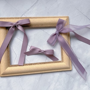 May include: Three lavender hair bows of different sizes are arranged on a gold picture frame. The bows are made of satin and sheer ribbon. The frame is rectangular with a decorative edge. The bows are a soft, muted purple colour.