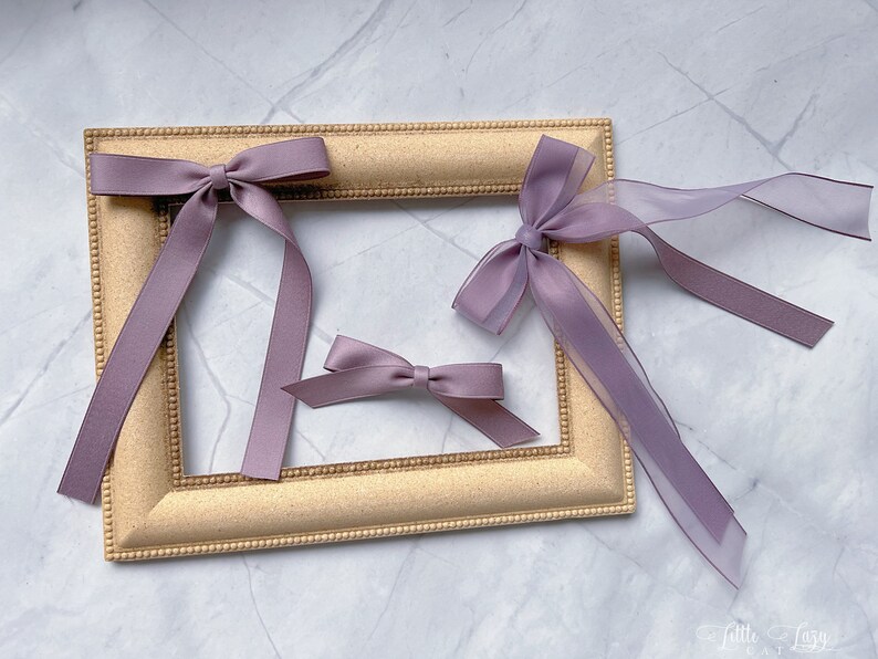 May include: Three lavender hair bows of different sizes are arranged on a gold picture frame. The bows are made of satin and sheer ribbon. The frame is rectangular with a decorative edge. The bows are a soft, muted purple colour.