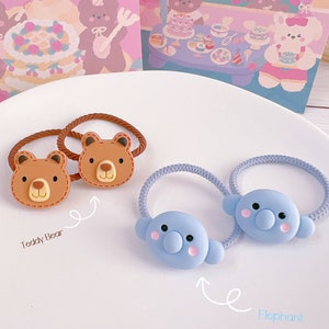 May include: Two hair ties with brown teddy bear faces and two with blue elephant faces. The teddy bear ties have brown elastic bands, while the elephant ties have blue elastic bands. The background includes two illustrated cards.