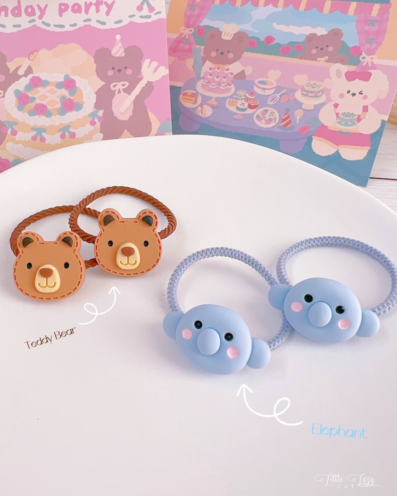 May include: Two hair ties with brown teddy bear faces and two with blue elephant faces. The teddy bear ties have brown elastic bands, while the elephant ties have blue elastic bands. The background includes two illustrated cards.