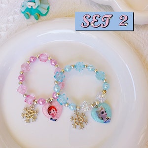 K&ouml;nnte beinhalten: Zwei Armb&auml;nder mit Perlen und herzf&ouml;rmigen Anh&auml;ngern mit Zeichentrickfiguren und Schneeflockenanh&auml;ngern. Ein Armband ist rosa, das andere blau. Die Armb&auml;nder liegen auf einem wei&szlig;en Teller, mit dem Text "SET 2" in einer hellblauen Box.