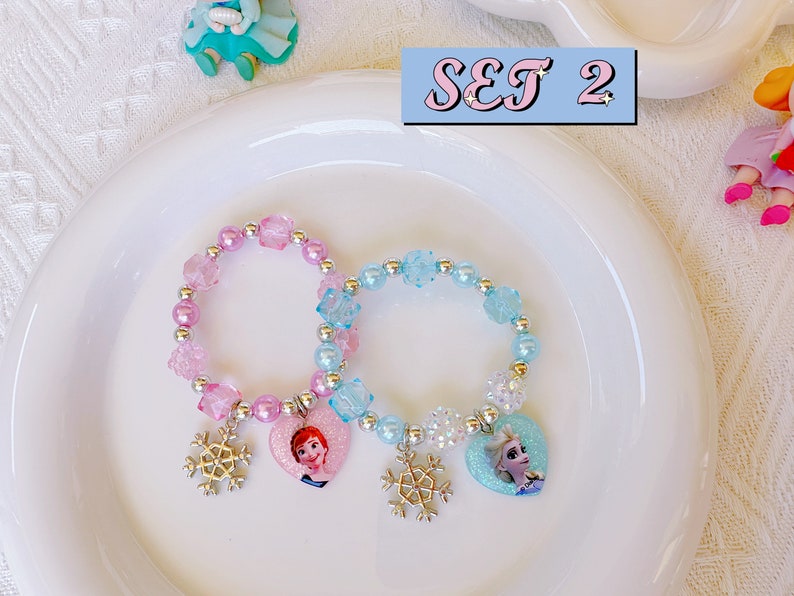 K&ouml;nnte beinhalten: Zwei Armb&auml;nder mit Perlen und herzf&ouml;rmigen Anh&auml;ngern mit Zeichentrickfiguren und Schneeflockenanh&auml;ngern. Ein Armband ist rosa, das andere blau. Die Armb&auml;nder liegen auf einem wei&szlig;en Teller, mit dem Text "SET 2" in einer hellblauen Box.