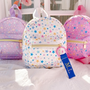 Puede incluir: Tres pequeñas mochilas acolchadas con estampados de estrellas en colores pastel. La mochila central es blanca con estrellas azules, amarillas y rosas. Las otras dos son lavanda y rosa. Cada una tiene una cremallera dorada y un asa superior.