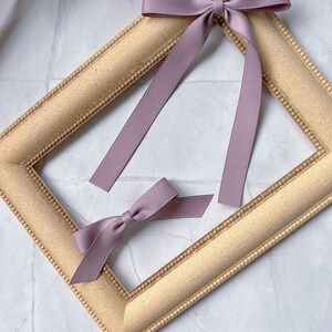 May include: Two mauve ribbon bows, one tied with long streamers, the other smaller and untied. They rest on a gold-coloured, rectangular picture frame with a beaded edge. The background is a white marble surface.