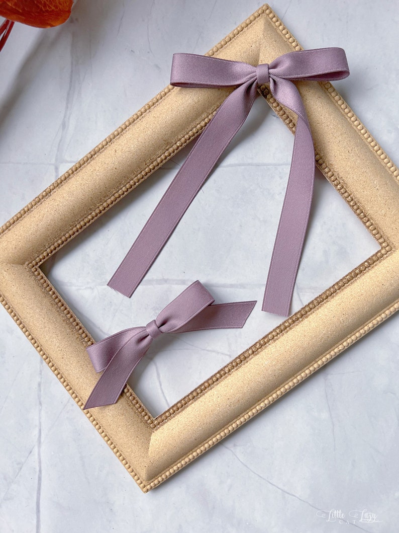 May include: Two mauve ribbon bows, one tied with long streamers, the other smaller and untied. They rest on a gold-coloured, rectangular picture frame with a beaded edge. The background is a white marble surface.