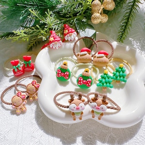 Puede incluir: Una colección de coleteros con temática navideña, con varios diseños, incluyendo gorros de Papá Noel, árboles de Navidad, adornos, renos, hombrecitos de jengibre y figuras de gnomos. Los coleteros están dispuestos en un plato blanco con borde festoneado, con follaje verde en el fondo.