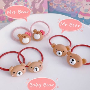 May include: A collection of brown bear-shaped hair ties with red and pink elastic bands. The bear faces have white and black details, with some bears wearing pink bows. The image includes text bubbles that say "Mrs Bear", "Mr Bear", and "Baby Bear".