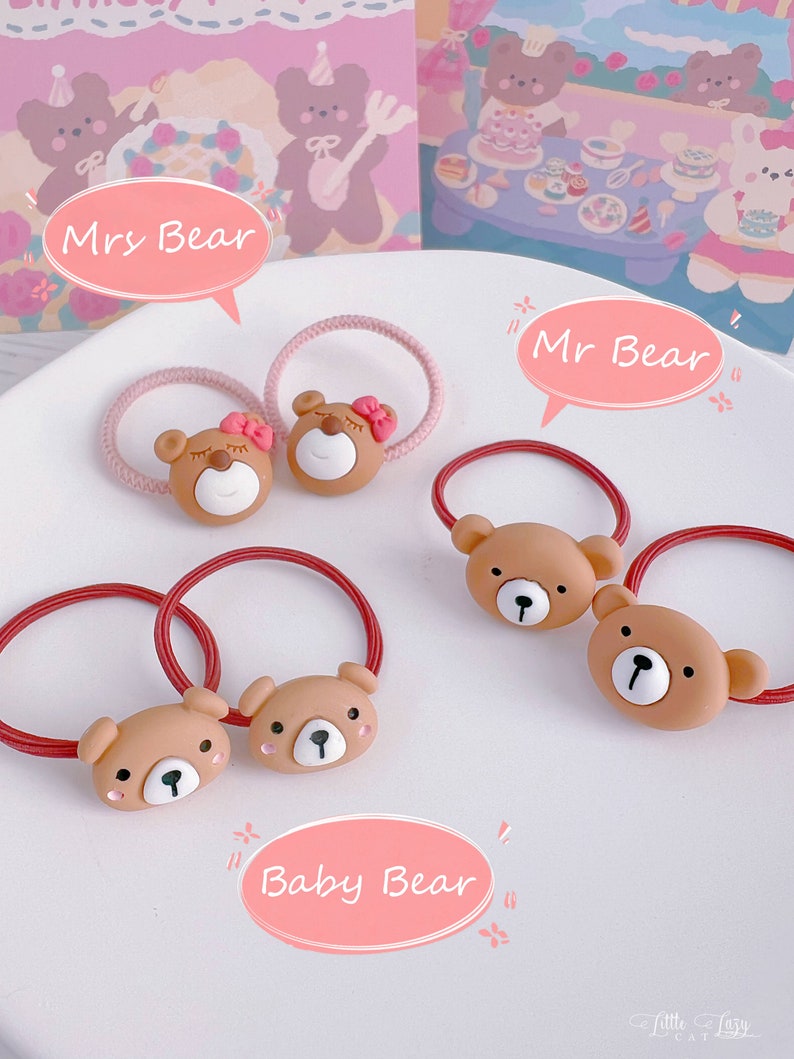 May include: A collection of brown bear-shaped hair ties with red and pink elastic bands. The bear faces have white and black details, with some bears wearing pink bows. The image includes text bubbles that say "Mrs Bear", "Mr Bear", and "Baby Bear".