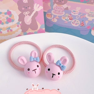 May include: Two pink bunny hair ties with blue bows and black facial features. The hair ties are on a pink elastic band. The image also includes a pink card with a bear illustration and the text "Cute Bunny".