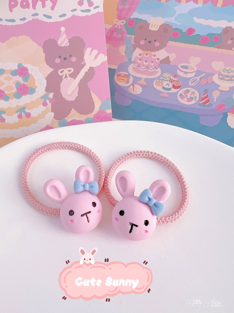 May include: Two pink bunny hair ties with blue bows and black facial features. The hair ties are on a pink elastic band. The image also includes a pink card with a bear illustration and the text "Cute Bunny".