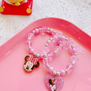 May include: Two pink beaded bracelets with Minnie Mouse charms. One bracelet features a Minnie Mouse head charm, the other has a heart-shaped charm with a Minnie Mouse design. The bracelets are on a pink tray.