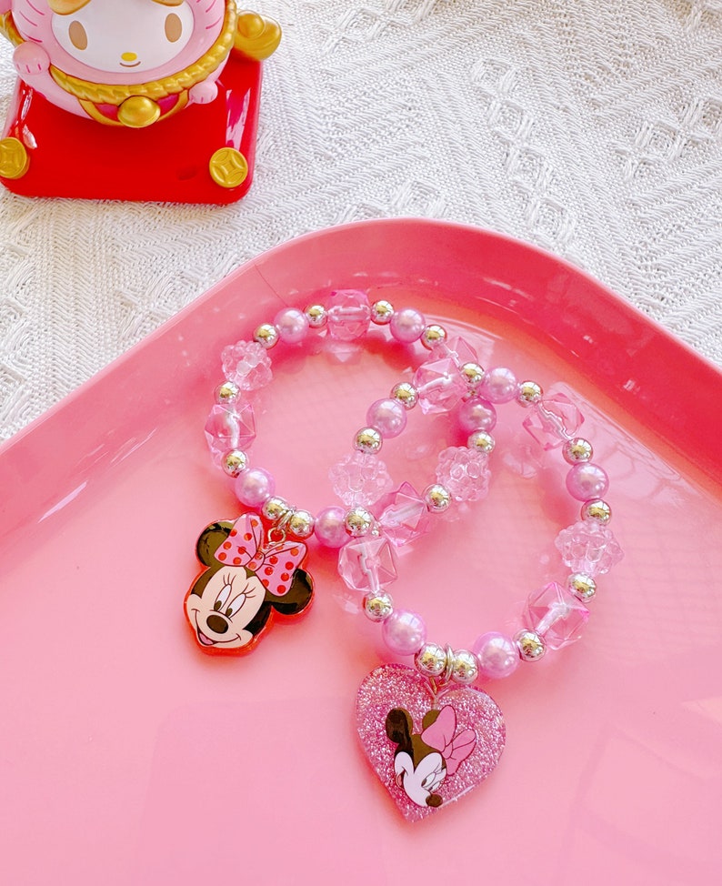 May include: Two pink beaded bracelets with Minnie Mouse charms. One bracelet features a Minnie Mouse head charm, the other has a heart-shaped charm with a Minnie Mouse design. The bracelets are on a pink tray.