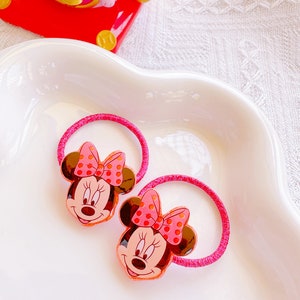 May include: Two pink Minnie Mouse hair ties with brown ears and a pink polka dot bow. The hair ties are on a white, wavy-edged dish. The text "Minnie 2" is at the bottom of the image.