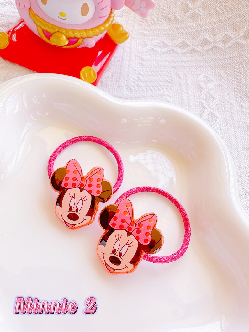 May include: Two pink Minnie Mouse hair ties with brown ears and a pink polka dot bow. The hair ties are on a white, wavy-edged dish. The text "Minnie 2" is at the bottom of the image.