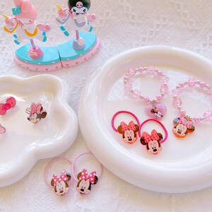 May include: A collection of pink accessories, including bracelets, hair ties, and rings, featuring Minnie Mouse and bow designs. The items are arranged on white, decorative plates, with a decorative figurine in the background.