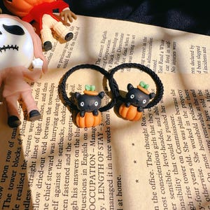Pode incluir: Duas presilhas de cabelo pretas com pingentes de Halloween. Cada pingente apresenta um gato preto com um chap&eacute;u verde, sentado em uma ab&oacute;bora laranja. Um rosto de Jack-o'-lantern e texto de livro no fundo.