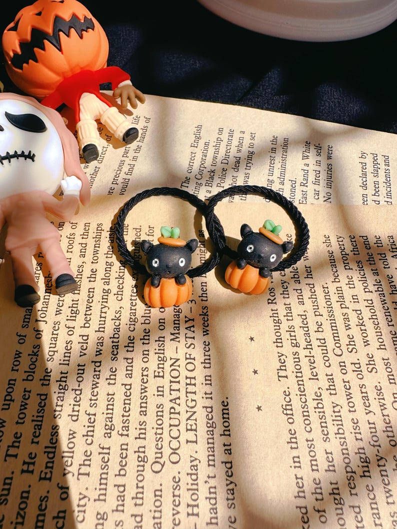 Pode incluir: Duas presilhas de cabelo pretas com pingentes de Halloween. Cada pingente apresenta um gato preto com um chap&eacute;u verde, sentado em uma ab&oacute;bora laranja. Um rosto de Jack-o'-lantern e texto de livro no fundo.