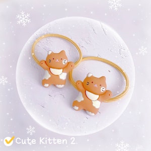 Puede incluir: Dos gomas para el pelo con figuras de gatitos marrones y blancos. Las figuras de gatitos tienen los brazos extendidos y est&aacute;n unidas a una banda el&aacute;stica amarilla. El texto "Cute Kitten 2." es visible en la esquina inferior izquierda.