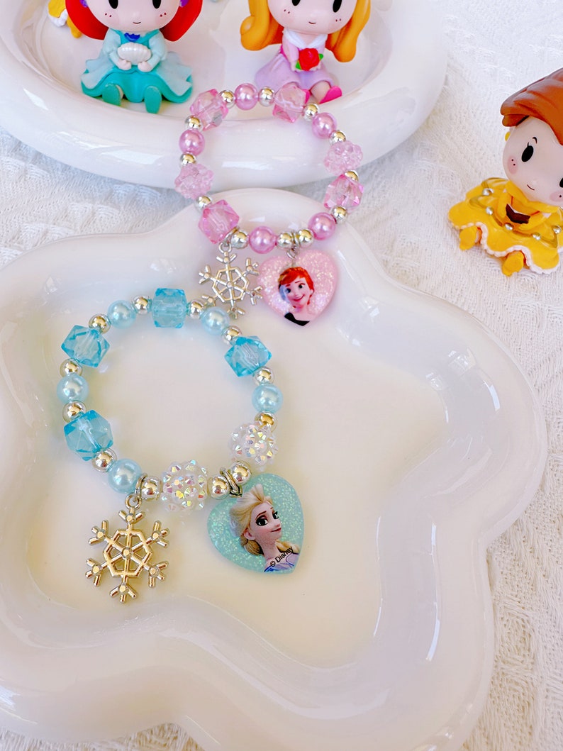 K&ouml;nnte beinhalten: Zwei Armb&auml;nder mit Perlen, Schneeflockenanh&auml;ngern und herzf&ouml;rmigen Anh&auml;ngern mit Zeichentrickfiguren. Ein Armband ist rosa mit einem Herzanh&auml;nger einer Figur, das andere ist blau. Dekorative Figuren im Hintergrund.