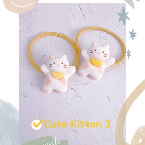 Puede incluir: Dos gomas para el pelo blancas con forma de gatito, con detalles rosas y cuellos amarillos. Las gomas tienen una banda el&aacute;stica amarilla. El texto "Cute Kitten 3" es visible en amarillo. El fondo tiene un dise&ntilde;o azul claro y blanco.