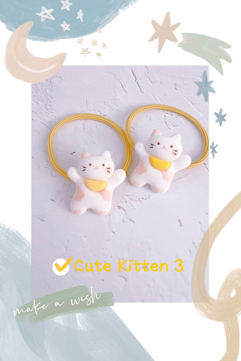 Puede incluir: Dos gomas para el pelo blancas con forma de gatito, con detalles rosas y cuellos amarillos. Las gomas tienen una banda el&aacute;stica amarilla. El texto "Cute Kitten 3" es visible en amarillo. El fondo tiene un dise&ntilde;o azul claro y blanco.