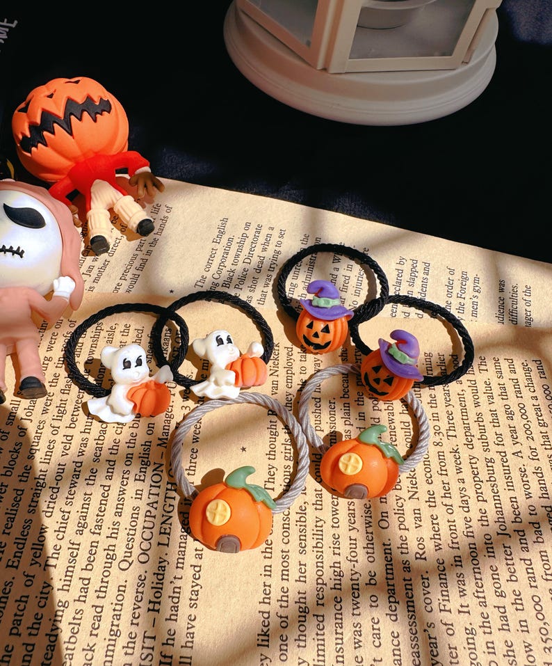 Pode incluir: Uma cole&ccedil;&atilde;o de el&aacute;sticos de cabelo com tema de Halloween. Os el&aacute;sticos apresentam ab&oacute;boras laranjas, fantasmas e jack-o'-lanterns. Alguns t&ecirc;m chap&eacute;us de bruxa. O fundo &eacute; um papel impresso.