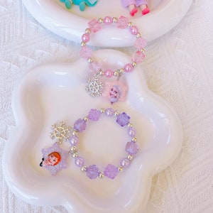 K&ouml;nnte beinhalten: Zwei Armb&auml;nder mit Perlen in einer wei&szlig;en Schale. Eines ist rosa mit einem Schneeflocken-Charm und einem Prinzessinnen-Anh&auml;nger. Das andere ist lila mit einem Schneeflocken-Charm und einem Prinzessinnen-Anh&auml;nger. Im Hintergrund sind zwei Prinzessinnenfiguren.