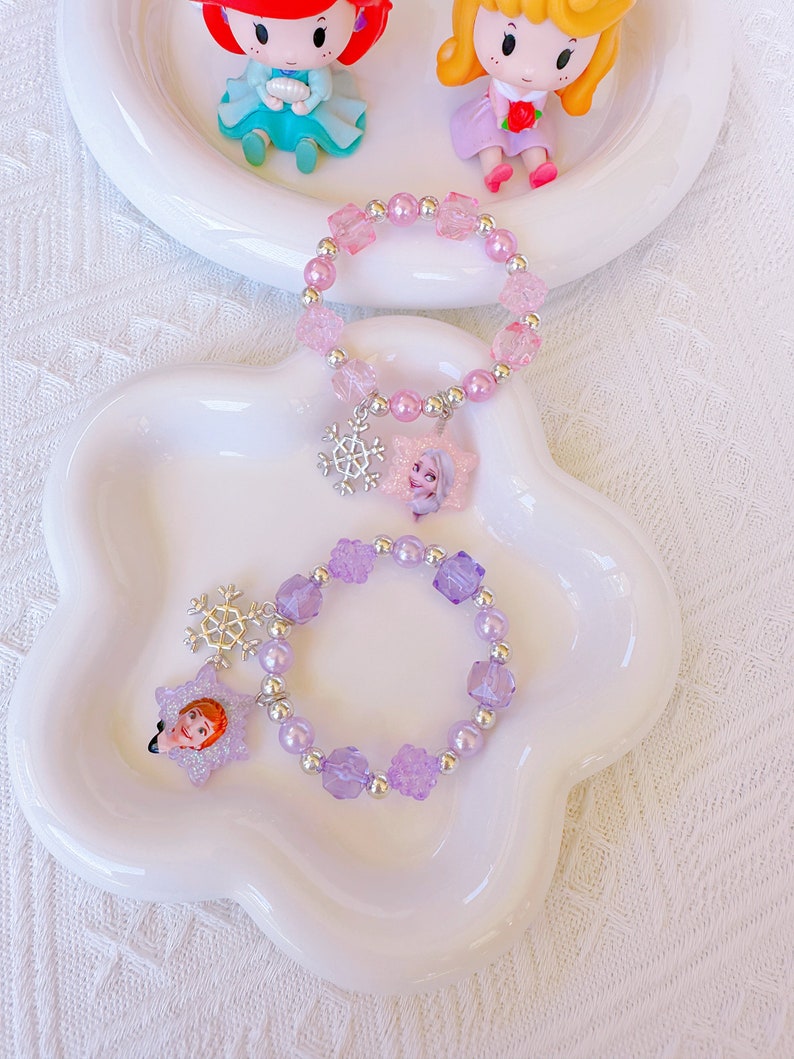 K&ouml;nnte beinhalten: Zwei Armb&auml;nder mit Perlen in einer wei&szlig;en Schale. Eines ist rosa mit einem Schneeflocken-Charm und einem Prinzessinnen-Anh&auml;nger. Das andere ist lila mit einem Schneeflocken-Charm und einem Prinzessinnen-Anh&auml;nger. Im Hintergrund sind zwei Prinzessinnenfiguren.