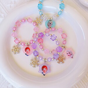K&ouml;nnte beinhalten: Eine Sammlung von Kinderschmuck, darunter Armb&auml;nder und eine Halskette, mit Winterthema. Die Armb&auml;nder und die Halskette sind mit bunten Perlen, Schneeflocken-Anh&auml;ngern und Charakter-Anh&auml;ngern verziert. Die Farbpalette umfasst Rosa, Lila und Blau.