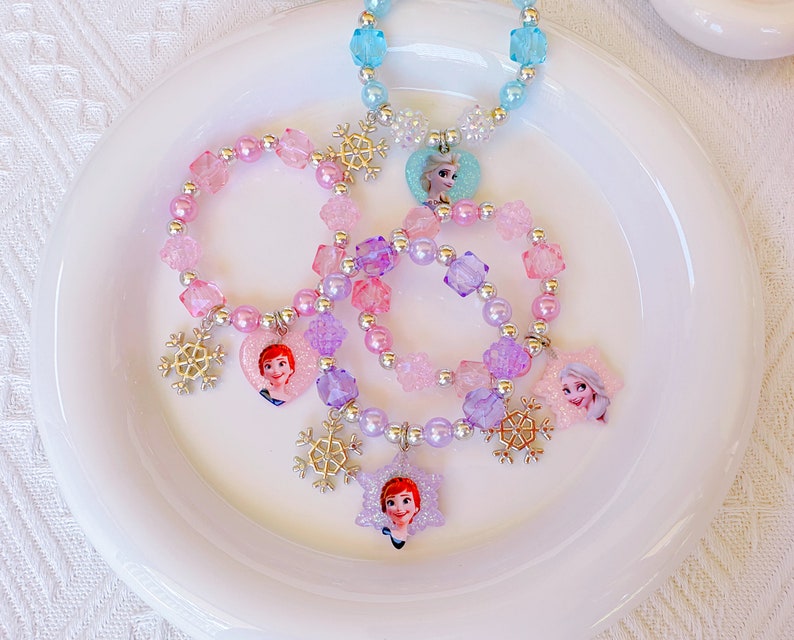K&ouml;nnte beinhalten: Eine Sammlung von Kinderschmuck, darunter Armb&auml;nder und eine Halskette, mit Winterthema. Die Armb&auml;nder und die Halskette sind mit bunten Perlen, Schneeflocken-Anh&auml;ngern und Charakter-Anh&auml;ngern verziert. Die Farbpalette umfasst Rosa, Lila und Blau.