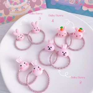 May include: A collection of pink hair ties, each adorned with a cute, cartoon-style bunny face. Some bunnies have carrot accents. The hair ties are arranged on a white plate, with the text "Baby Bunny" and numbers labeling each.