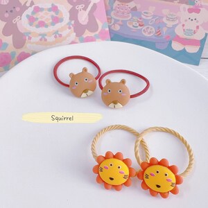 May include: Two sets of hair ties with animal designs. One set features brown squirrel faces on red bands, and the other has yellow lion faces with orange manes on tan bands. The word "Squirrel" and "Lion" are written below each set.