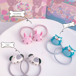 May include: A collection of hair ties with animal-themed designs. The ties feature pink bunny, blue owl, and white dog faces. The hair ties are on a white plate with a pink and white background. The text on the image says "Funny Bunny", "Owlet", and "Doggies".