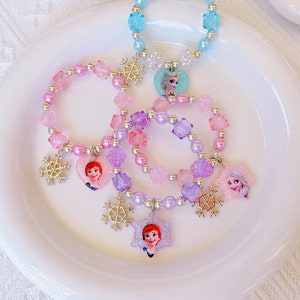 K&ouml;nnte beinhalten: Eine Sammlung von Kinderschmuck, darunter Armb&auml;nder und eine Halskette, mit bunten Perlen und Charakteranh&auml;ngern. Die Armb&auml;nder und die Halskette sind mit Schneeflocken-Anh&auml;ngern und Charakterbildern in Rosa-, Lila- und Blaut&ouml;nen verziert.
