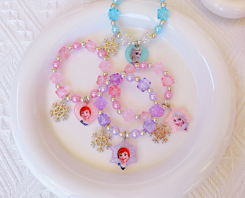 K&ouml;nnte beinhalten: Eine Sammlung von Kinderschmuck, darunter Armb&auml;nder und eine Halskette, mit bunten Perlen und Charakteranh&auml;ngern. Die Armb&auml;nder und die Halskette sind mit Schneeflocken-Anh&auml;ngern und Charakterbildern in Rosa-, Lila- und Blaut&ouml;nen verziert.