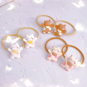 Puede incluir: Colecci&oacute;n de coleteros con adorables adornos en forma de gato en varios colores. Los adornos son blancos, marrones y rosas, con detalles amarillos y rosas. Las bandas el&aacute;sticas son de color amarillo claro.