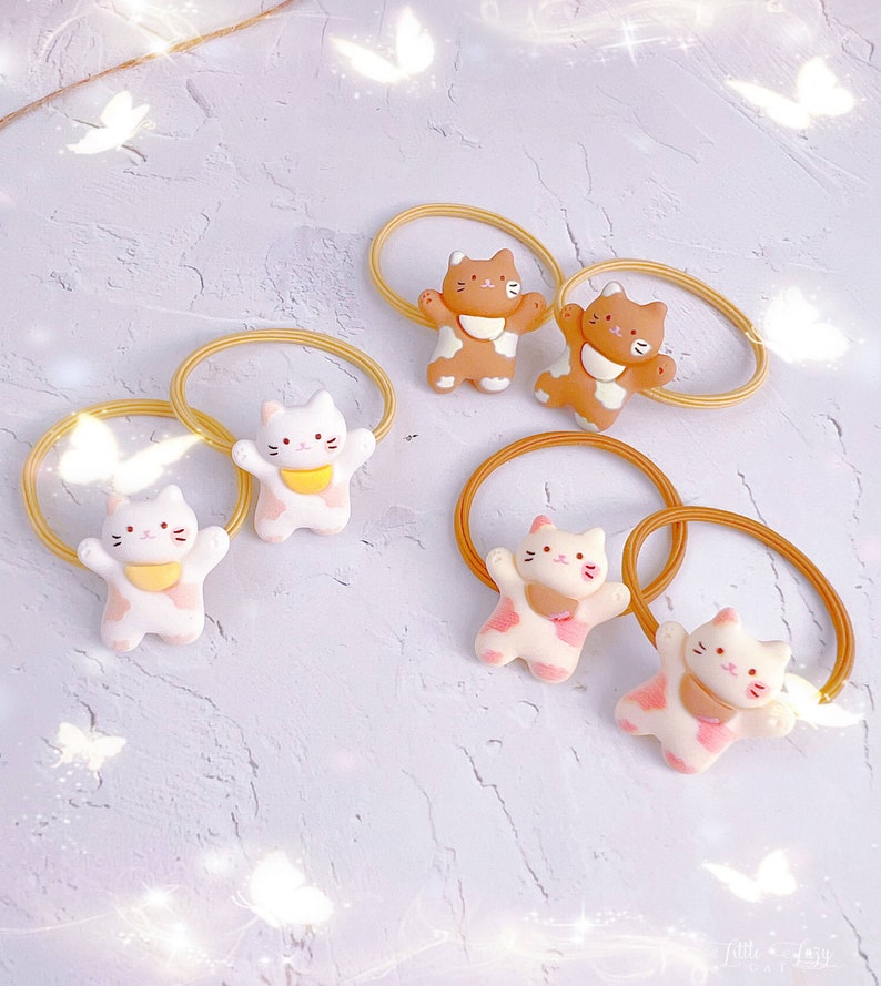 Puede incluir: Colecci&oacute;n de coleteros con adorables adornos en forma de gato en varios colores. Los adornos son blancos, marrones y rosas, con detalles amarillos y rosas. Las bandas el&aacute;sticas son de color amarillo claro.