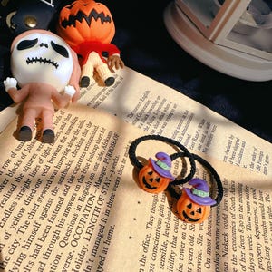 Pode incluir: El&aacute;sticos de cabelo com tema de Halloween com pingentes de ab&oacute;bora laranja com chap&eacute;us de bruxa roxos. A imagem tamb&eacute;m mostra estatuetas de personagens de O Estranho Mundo de Jack.