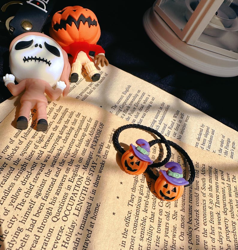Pode incluir: El&aacute;sticos de cabelo com tema de Halloween com pingentes de ab&oacute;bora laranja com chap&eacute;us de bruxa roxos. A imagem tamb&eacute;m mostra estatuetas de personagens de O Estranho Mundo de Jack.