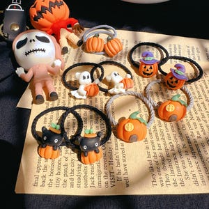 Pode incluir: Uma cole&ccedil;&atilde;o de el&aacute;sticos de cabelo e figuras com tema de Halloween. Os el&aacute;sticos de cabelo apresentam desenhos de ab&oacute;bora, fantasma e gato preto. Outros itens incluem uma figura com cabe&ccedil;a de ab&oacute;bora e uma figura de esqueleto. O fundo inclui texto impresso.