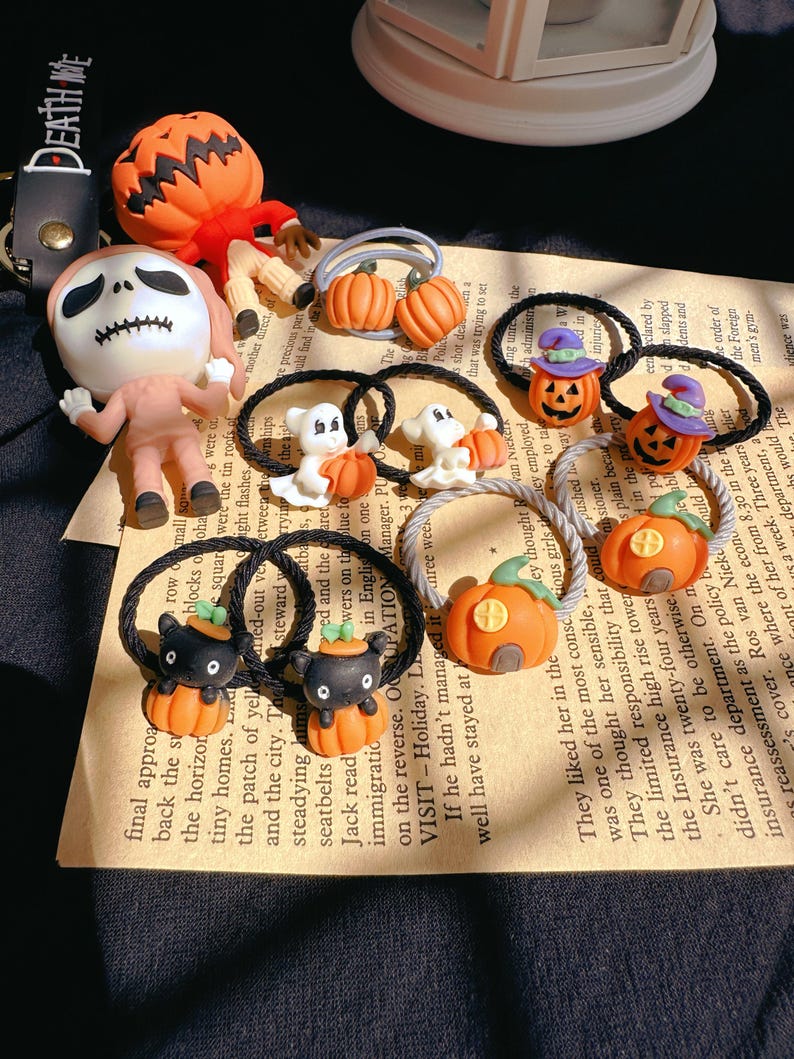Pode incluir: Uma cole&ccedil;&atilde;o de el&aacute;sticos de cabelo e figuras com tema de Halloween. Os el&aacute;sticos de cabelo apresentam desenhos de ab&oacute;bora, fantasma e gato preto. Outros itens incluem uma figura com cabe&ccedil;a de ab&oacute;bora e uma figura de esqueleto. O fundo inclui texto impresso.