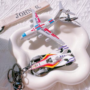 May include: A white airplane and race car keychain set with red, blue, and yellow accents. The airplane has the text "SUPER AVIATE" and the race car has the text "AD 400". The set is on a white, flower-shaped dish.