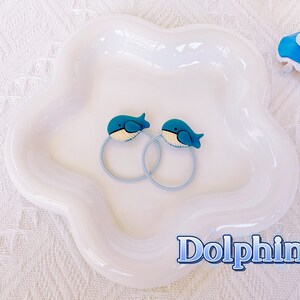 May include: Two blue whale-shaped hair ties with light blue bands rest on a white, flower-shaped dish. The whale designs have white bellies and are set against a white background. The word "Dolphin" is written in blue.
