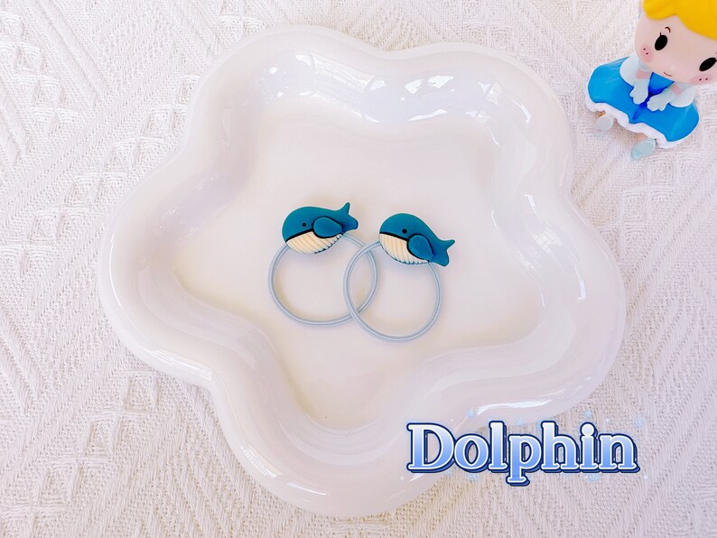 May include: Two blue whale-shaped hair ties with light blue bands rest on a white, flower-shaped dish. The whale designs have white bellies and are set against a white background. The word "Dolphin" is written in blue.