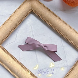 May include: A mauve ribbon bow, labelled 'Style 2', sits within a gold-coloured picture frame. The frame is on a white and grey marble-patterned surface. The bow has a simple design with two long, angled tails.