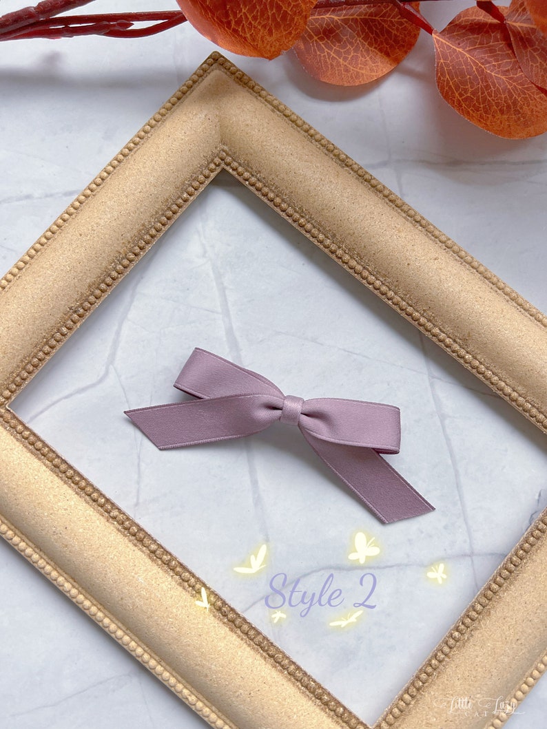 May include: A mauve ribbon bow, labelled 'Style 2', sits within a gold-coloured picture frame. The frame is on a white and grey marble-patterned surface. The bow has a simple design with two long, angled tails.
