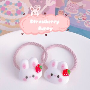May include: Two white bunny hair ties with pink cheeks and a red strawberry accent. The hair ties have a pink rope-like band. The image includes a pink cloud-shaped sign that says "Strawberry Bunny".
